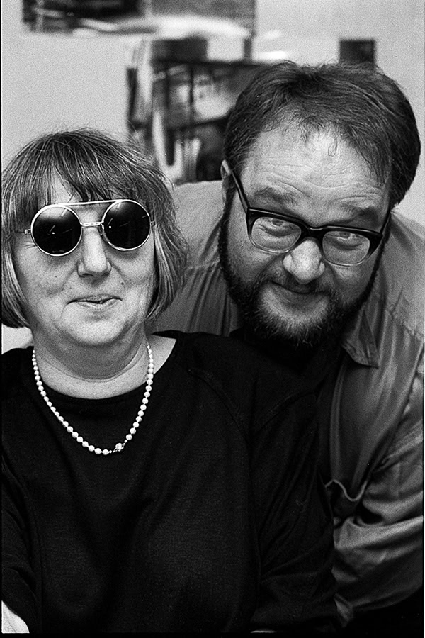Eine Frau mit grau meliertem Bob und Sonnenbrille und ein Mann mit Vollbart und Brille schauen beide verschmitzt in die Kamera