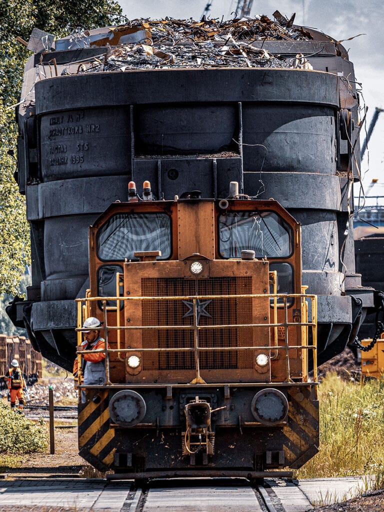 Frontalaufnahme einer gelb-orangen Rangierlokomotive auf einem Werksgleis. Ein Mitarbeiter mit weißem Helm und oranger Warnschutzkleidung sitzt auf dem seitlichen Umlauf am Führerhaus. Direkt dahinter ragt ein riesiger, schwarz verrußter Schrottkorb auf einem Waggon auf, oben quellen Schrottteile über den Rand. Eingeprägte Beschriftung am Korb: Inhalt 75 m³, Schrottkorb Nr. 2, Baujahr 1995. Links im Hintergrund ein zweiter Mitarbeiter in Warnkleidung.