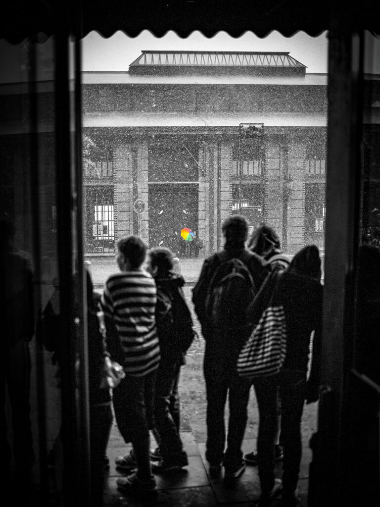Schwarz-weißes Streetfotografie-Bild aus einem Hauseingang heraus fotografiert: Mehrere Personen stehen im Rahmen einer Tordurchfahrt und blicken bei Regen auf ein gegenüberliegendes historisches Gebäude, der Hamburger Fischauktionshalle, mit Säulenarkaden und Glasoberlicht. Einzig ein bunter Regenschirm im Hintergrund ist farbig hervorgehoben, der Rest des Bilds ist monochrom.