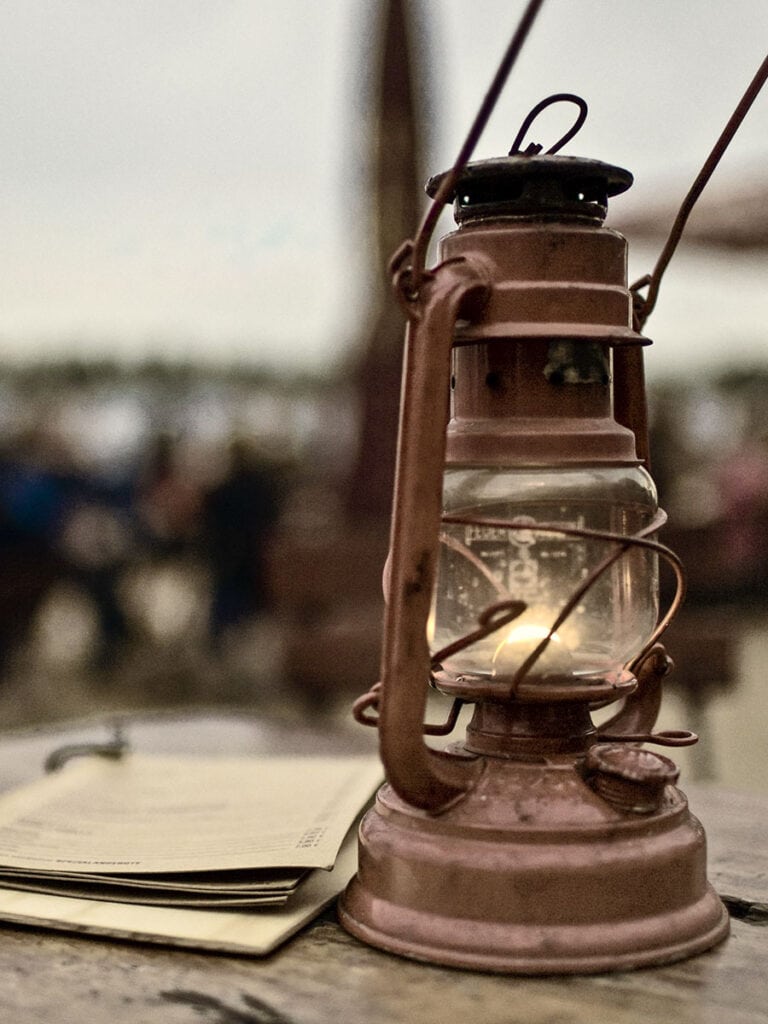 Eine brennende Sturmlaterne aus Metall auf einem Holztisch im Freien neben einer Speisekarte. Im Hintergrund unscharf Menschen an anderen Tischen und die Elbe.