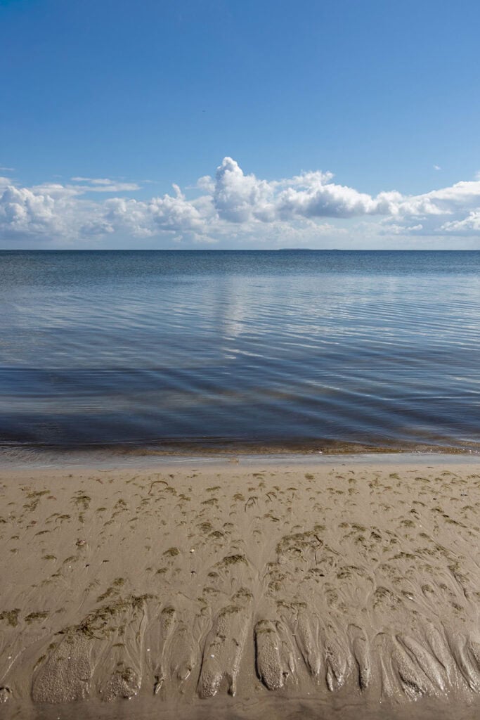 Blick vom Strand auf die ruhige Ostsee, in deren sanften Wellen sich die weißen Wolken im Hintergrund spiegeln. Der Himmel ist blau.