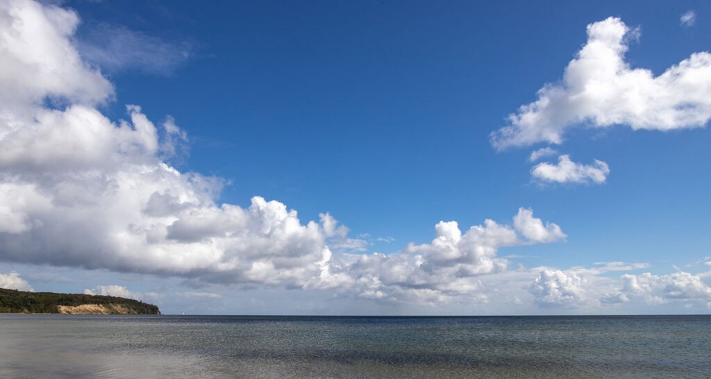Blick auf die Ostsee mit strahlend blauem Himmel und weißen Wolken, ruhiges Wasser, Küstenlinie mit Kreidefelsen im Hintergrund.