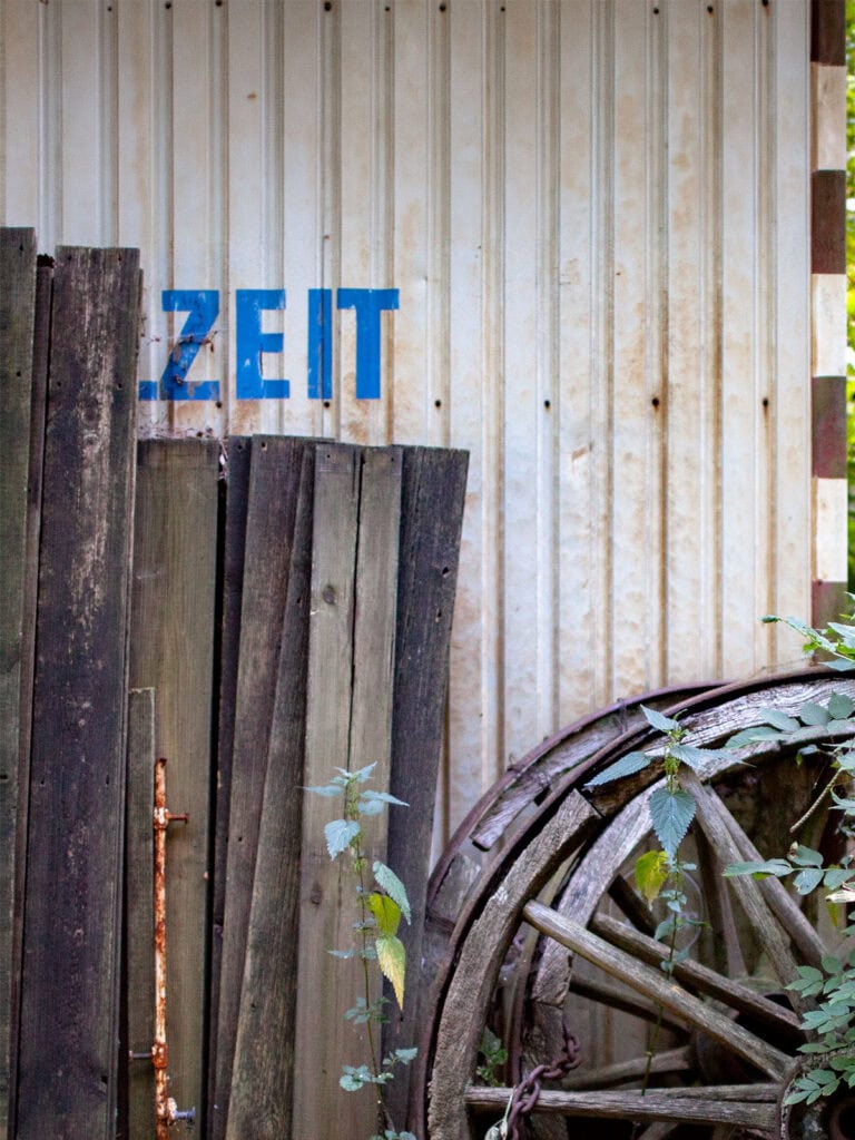 Alte verwitternde Holzräder und Holzpfosten vor einem Bauwagen mit dem Wort 'ZEIT' in Blau auf der Metallwand.