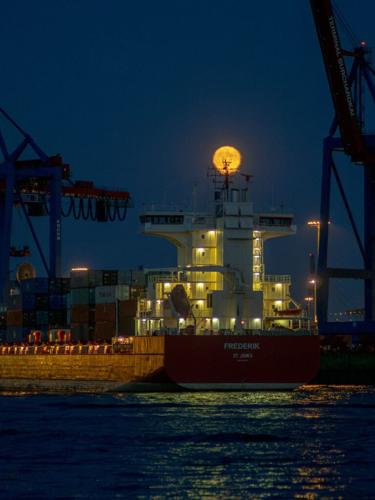 Vollmond über einem Hafen mit Containerschiffen und Kränen bei Nacht.