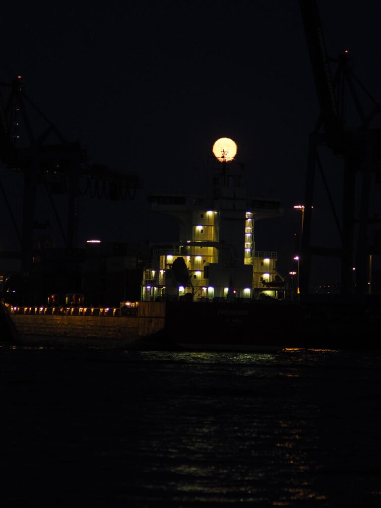 Vollmond über einem Hafen mit Kränen und beleuchteten Gebäuden bei Nacht.