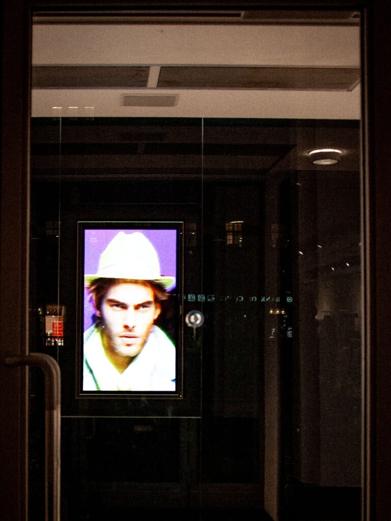 Schaufenster in der Nacht, das Modegeschäft liegt dunkel mit sanfter Notbeleuchtung im Hintergrund. Nur ein Hochklant-Monitor spielt farbenfrohe helle Fashion-Bilder ab. Hier sieht man ein männliches Model in weißer Kleidung und weißem Hut vor violettem Hintergrund.
