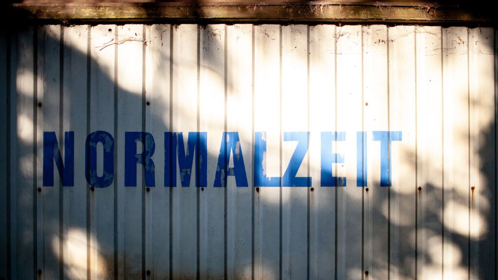 Blaue Normalzeit-Schrift auf der Metallwand eines alten Bauwagens mit Schatten.