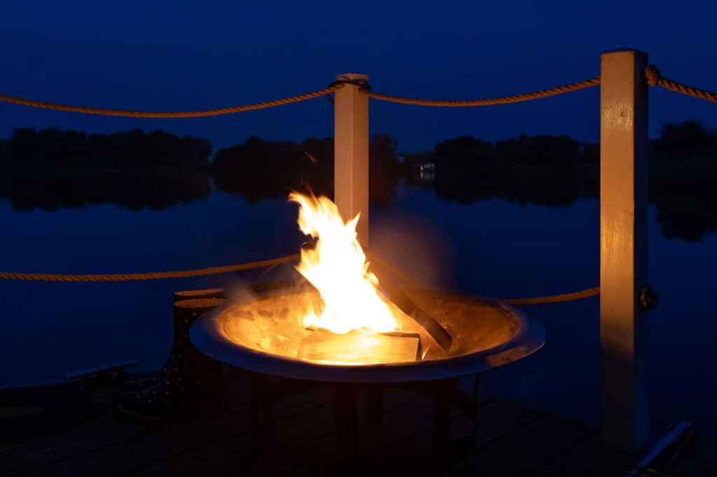 Lagerfeuer in einer Feuerschale auf einer Holzplattform bei Nacht am Wasser.